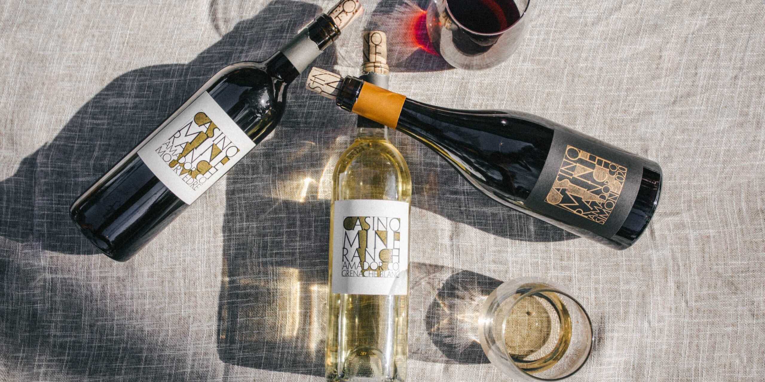 Three wine bottles, two red and one white, with labels showing "Casino Mine Ranch," are arranged on a textured surface with two glasses, one filled with red wine.