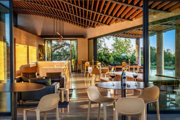 Modern restaurant interior with wooden ceiling, glass walls, and mixed seating. Tables are set with glasses and plates. Large windows offer a view of greenery outside.
