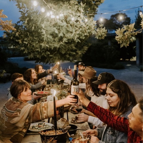 A group of people sitting at a long outdoor table, enjoying a meal and toasting with wine under string lights during the evening.
